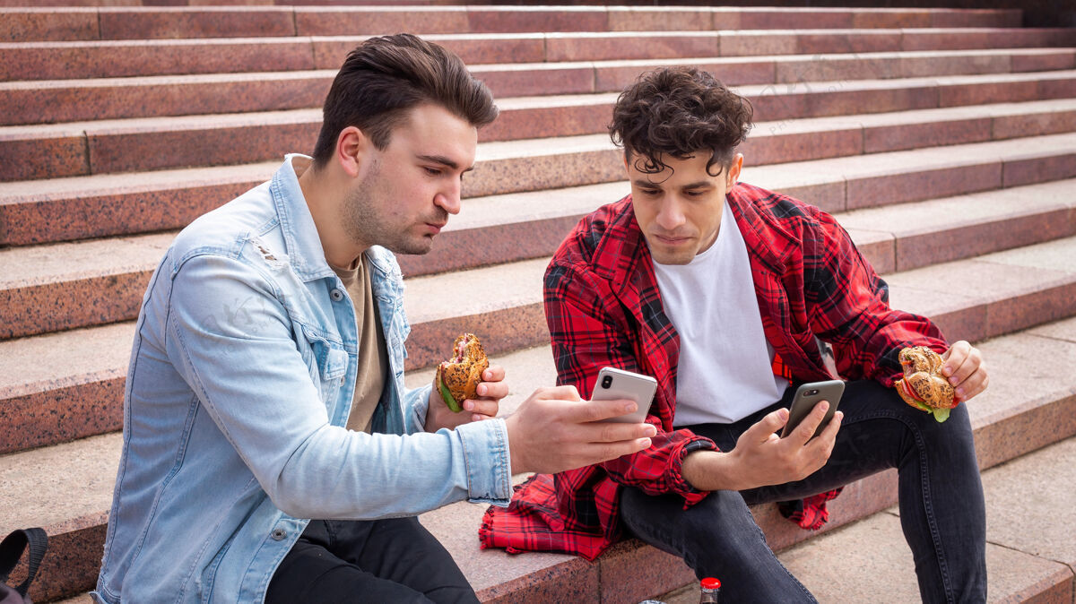帅哥两个年轻的朋友坐在公园的楼梯上吃饭城市生活方式可乐