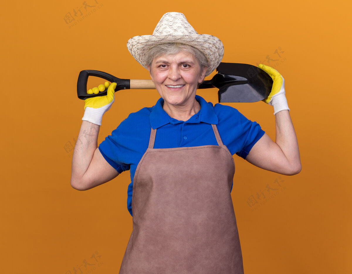 拿着面带微笑的上了年纪的女园丁戴着园艺帽和手套 背后脖子上拿着铲子园艺老人花园