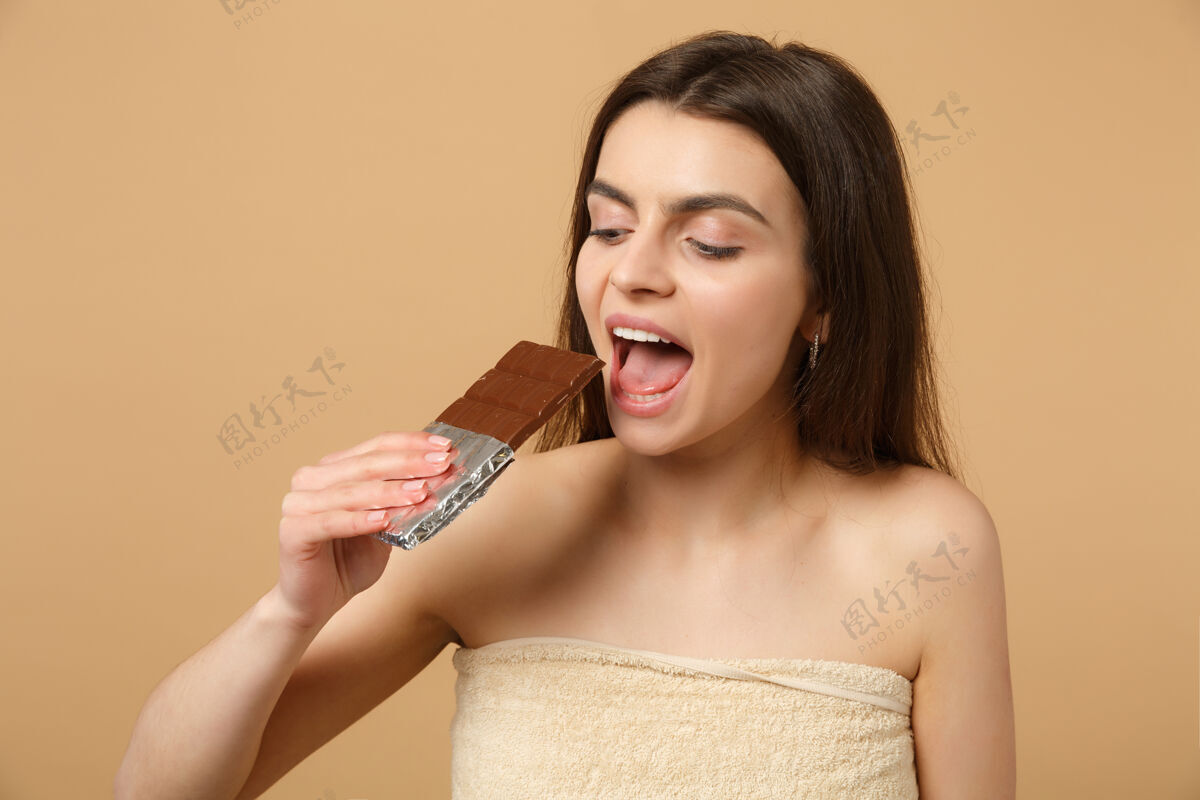 抗氧化剂近距离拍摄完美肌肤的半裸女人 裸妆将巧克力棒隔离在米色粉彩墙上女人咬糖果