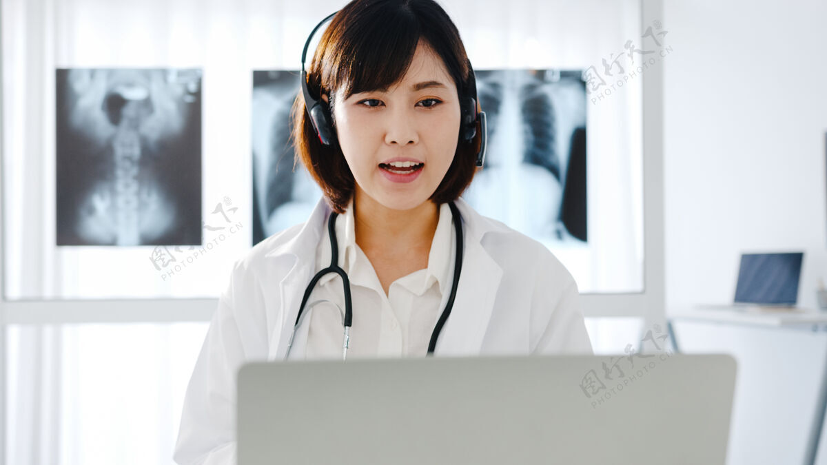 住宅年轻的亚洲女医生 身穿白色医疗制服 手持听诊器 使用笔记本电脑 在诊所或医院的办公桌上与患者进行视频会议呼叫医生咨询