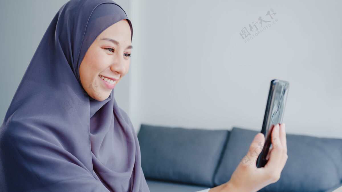 住宅年轻的亚洲穆斯林女商人使用智能手机通过视频聊天与朋友交谈头脑风暴在线会议 同时在客厅远程工作会议视频通话沙发