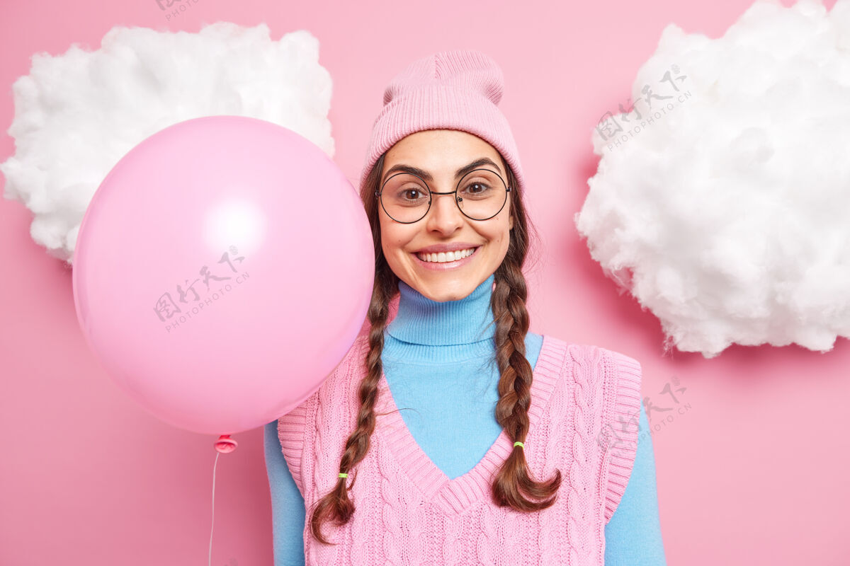 年轻女模特笑容可掬 穿着休闲服 留着两条梳好的长辫子 戴着圆眼镜 手持充气气球 期待节日或派对开始眼镜生日气球