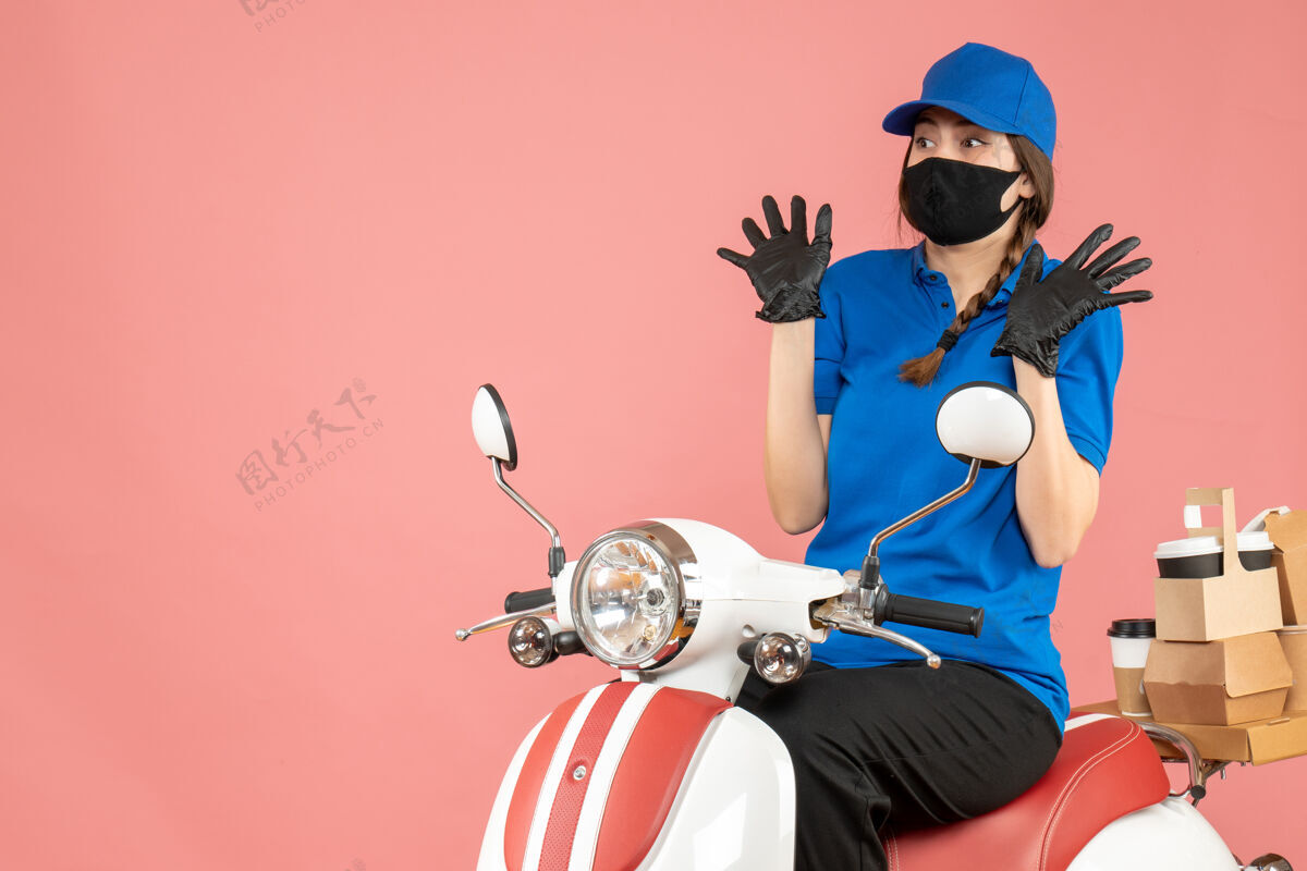 坐俯视图是一个戴着医用口罩和手套的女孩坐在滑板车上 在柔和的桃色背景下传递订单摩托车穿命令