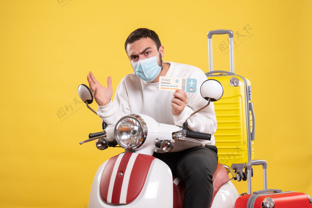 健身旅行概念的正面图 戴着医学面罩的年轻人坐在摩托车上 黄色手提箱 手里拿着车票人摩托车身体