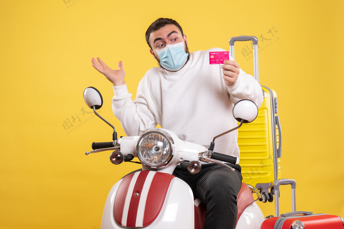 前面旅行概念的正面图 戴着医用面罩的年轻人坐在摩托车上 黄色手提箱 手里拿着银行卡面罩黄色人