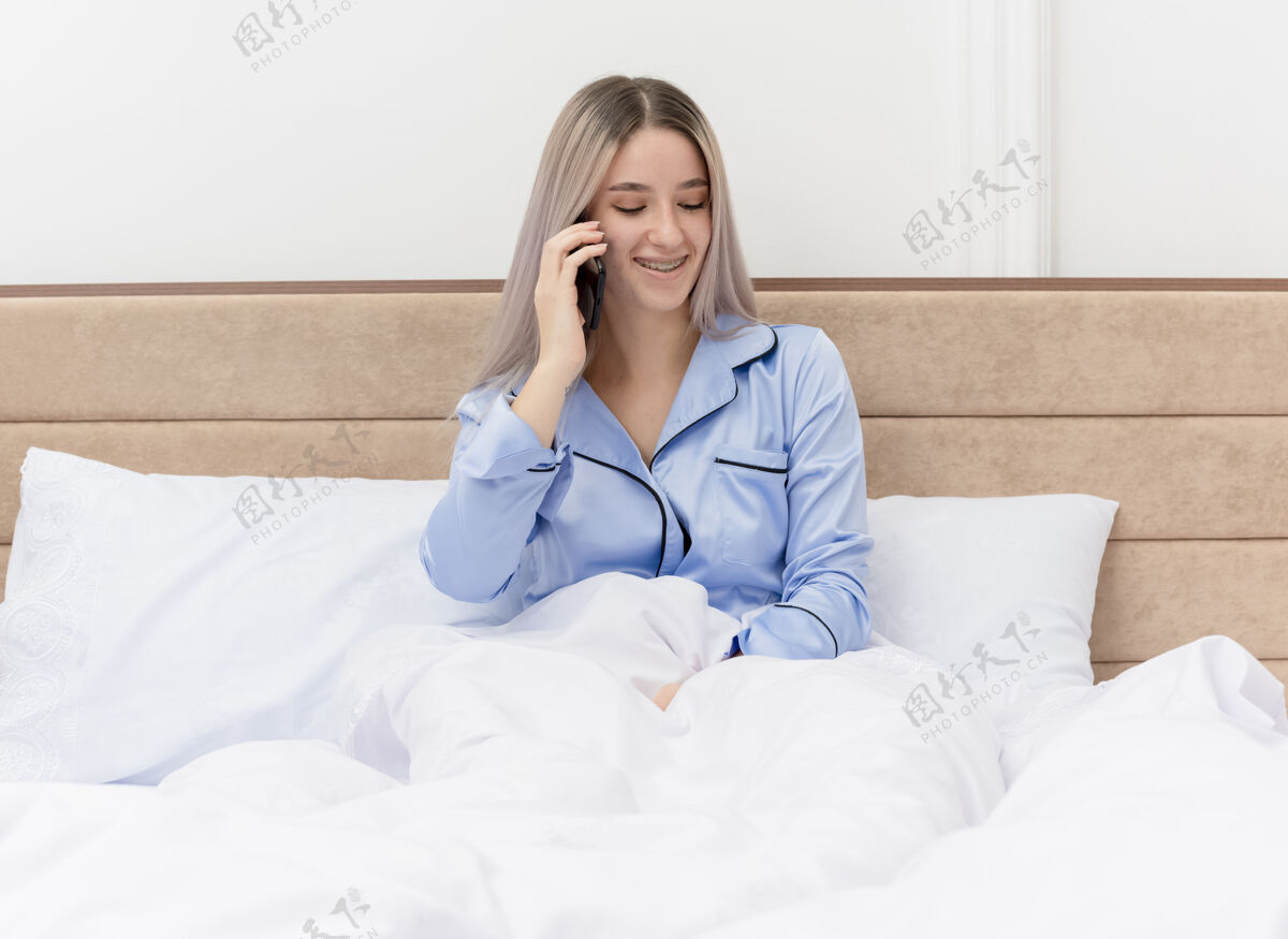 室内穿着蓝色睡衣的年轻美女坐在床上讲手机 在卧室里微笑女人卧室年轻