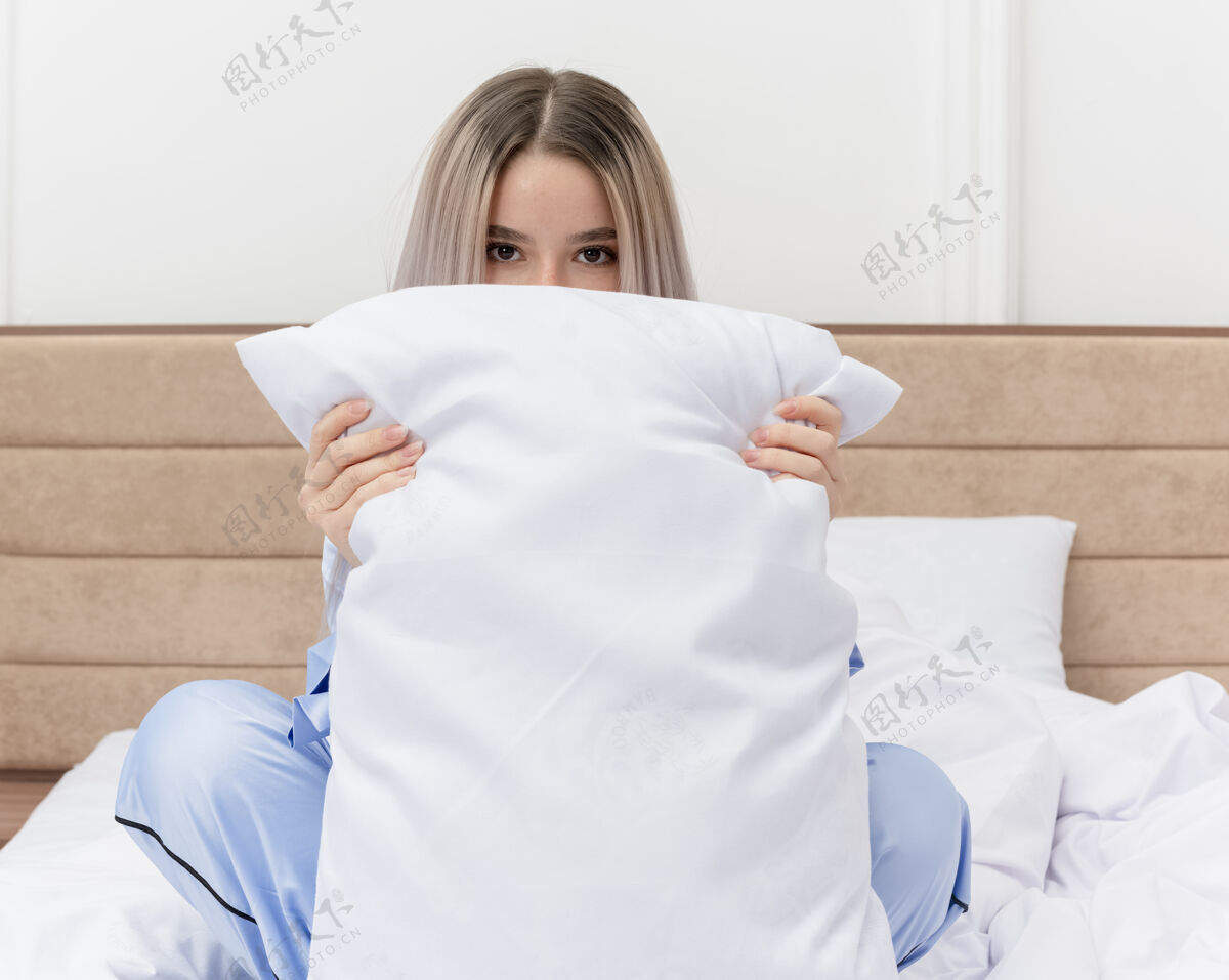枕头穿着蓝色睡衣的年轻漂亮女人坐在床上 枕头遮住了脸 在卧室里偷看漂亮卧室坐着