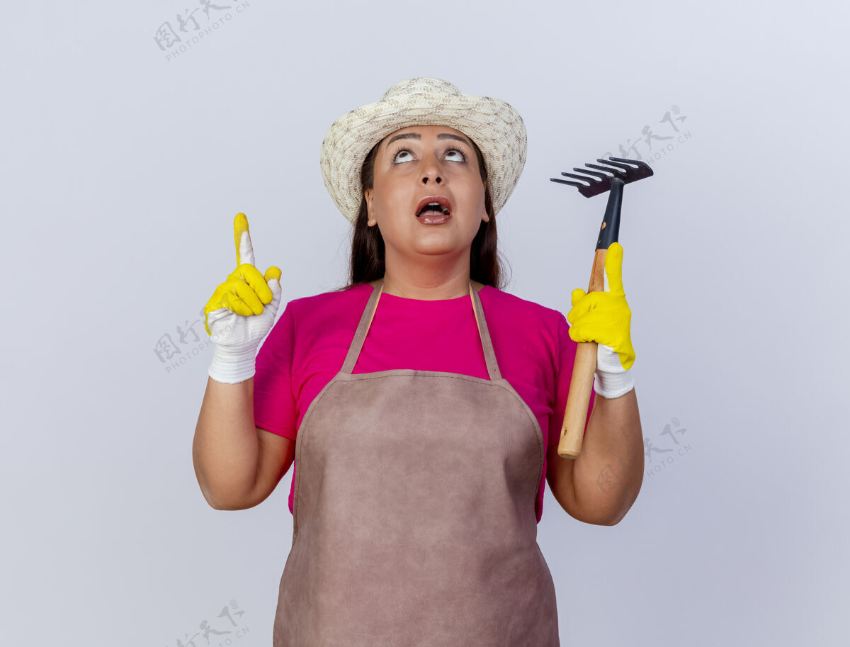 中围裙戴帽子的中年园丁妇女戴着橡胶手套拿着小耙子食指朝上指着惊讶花园穿上