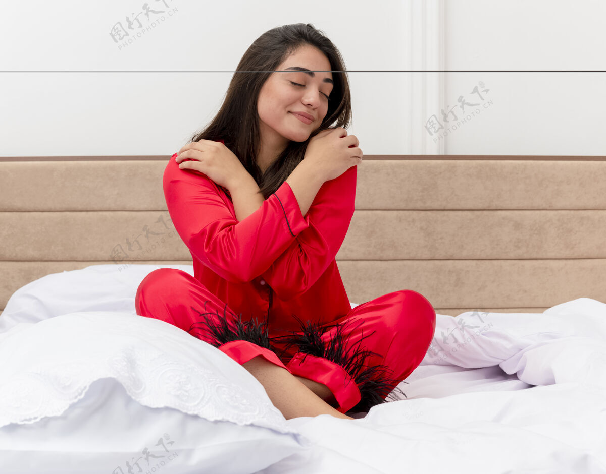 漂亮穿着红色睡衣的年轻漂亮女人坐在床上闭着眼睛拥抱着自己 在卧室里感受着积极的情绪她自己年轻眼睛