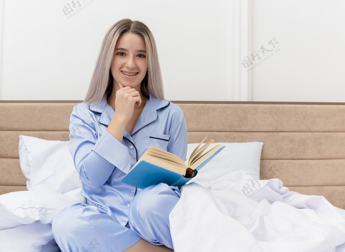 坐着穿着蓝色睡衣的年轻美女坐在床上 脸上挂着微笑的书在卧室里睡衣蓝色脸