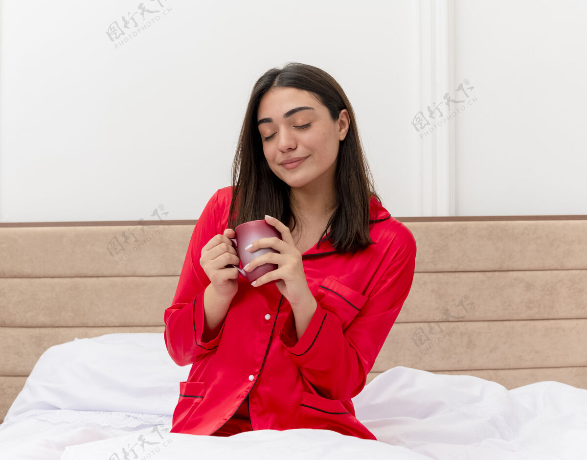 坐着穿着红色睡衣的年轻美女坐在床上 闭着眼睛喝着咖啡 感受卧室内部的积极情绪情绪杯子眼睛