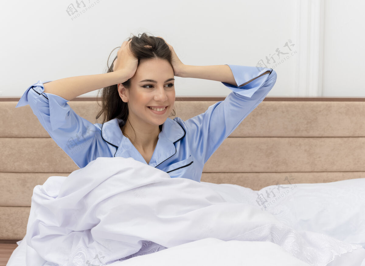 微笑穿着蓝色睡衣的年轻漂亮女人坐在床上快乐而积极地微笑着 愉快地抚摸着她在卧室里醒来的头室内女人背景