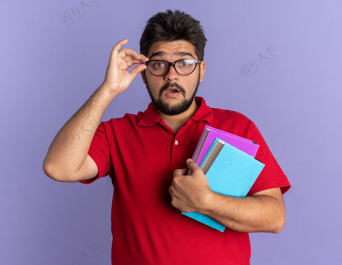 手势年轻的留着胡子的学生 穿着红色马球衫 戴着眼镜 手里拿着书 惊讶地站在蓝色的墙上马球胡须姿势