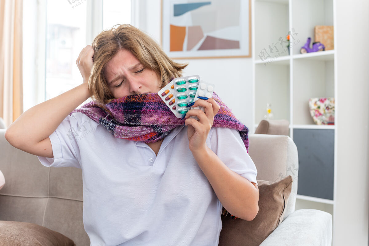 病不健康的年轻女子 脖子上围着暖和的围巾 感觉不舒服 生病 患流感 感冒 拿着不同的药片 坐在客厅的沙发上 看起来很担心沙发年轻流感