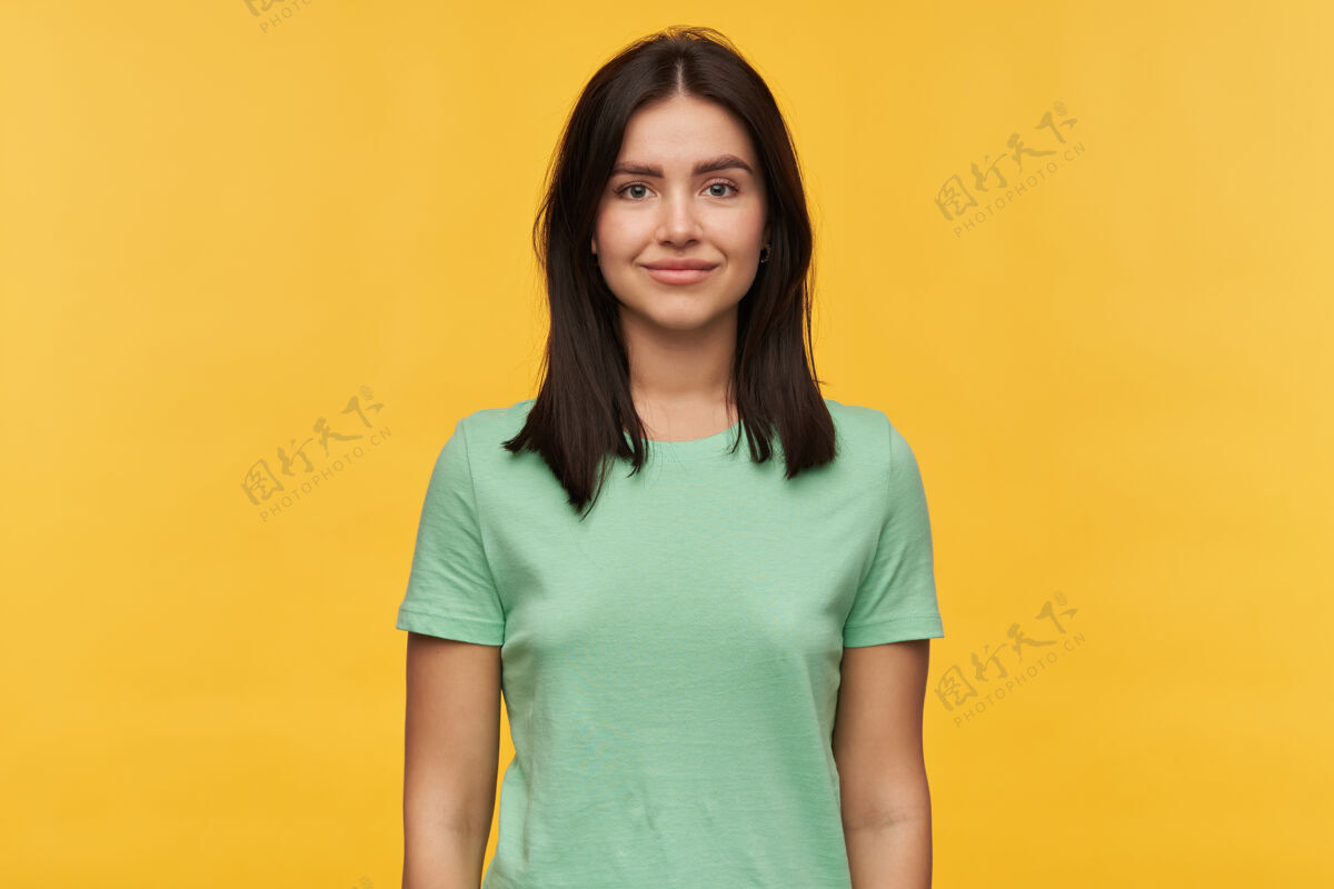 请身着薄荷色T恤的快乐美丽黑发年轻女子的肖像在黄色的墙壁上看起来很自信黑发女人姿势