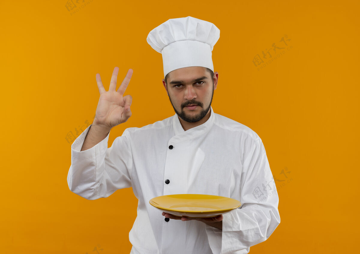 好的自信的年轻男性厨师 身着厨师制服 手拿空盘子 在橙色的墙上贴着“做得好”的牌子 上面留有复印空间盘子年轻烹饪