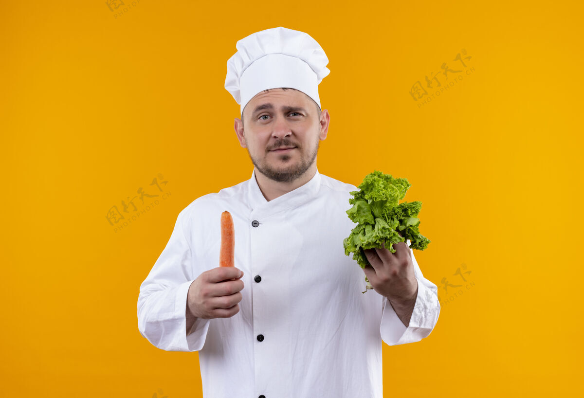 制服自信的年轻帅哥厨师穿着厨师制服拿着莴苣和胡萝卜隔离在橙色的墙上胡萝卜帅气厨师