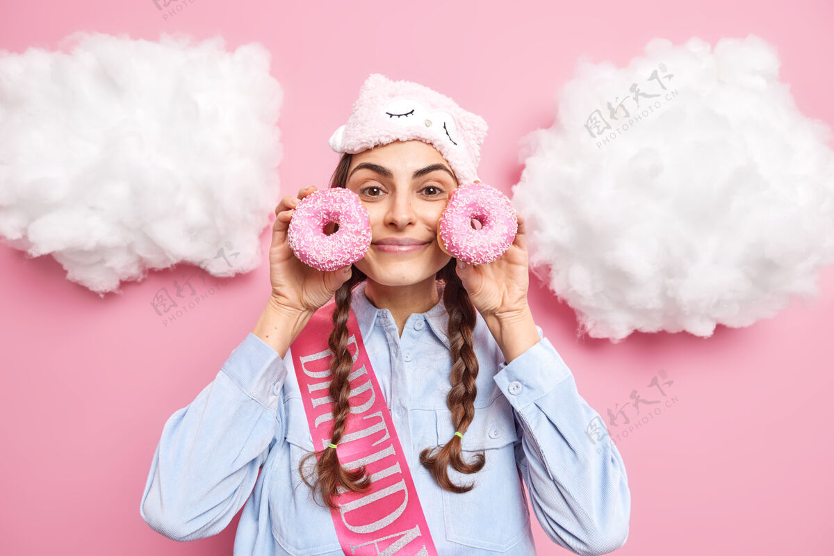 年轻扎着辫子的女人拿着玻璃甜甜圈在脸附近想吃面包店的产品在额头上戴上睡罩衬衫和丝带写着生日词在白云周围摆姿势快乐积极甜点