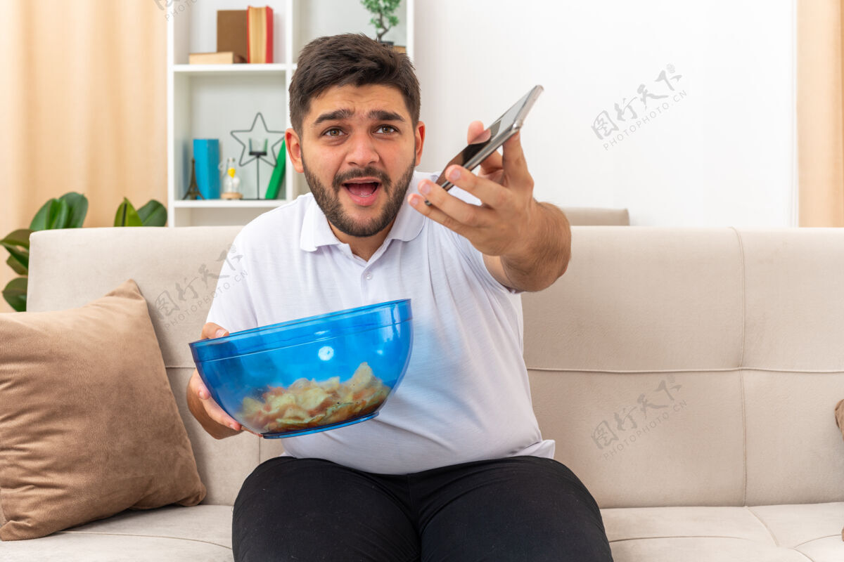 男人一个穿着休闲服的年轻人拿着一碗芯片和智能手机坐在客厅的沙发上 看起来很困惑生活碗芯片