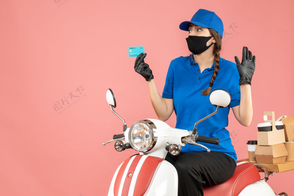 困惑前视图混乱的快递女孩戴着医用面罩和手套坐在滑板车上拿着银行卡在粉彩桃色背景下传递订单坐面具摩托车