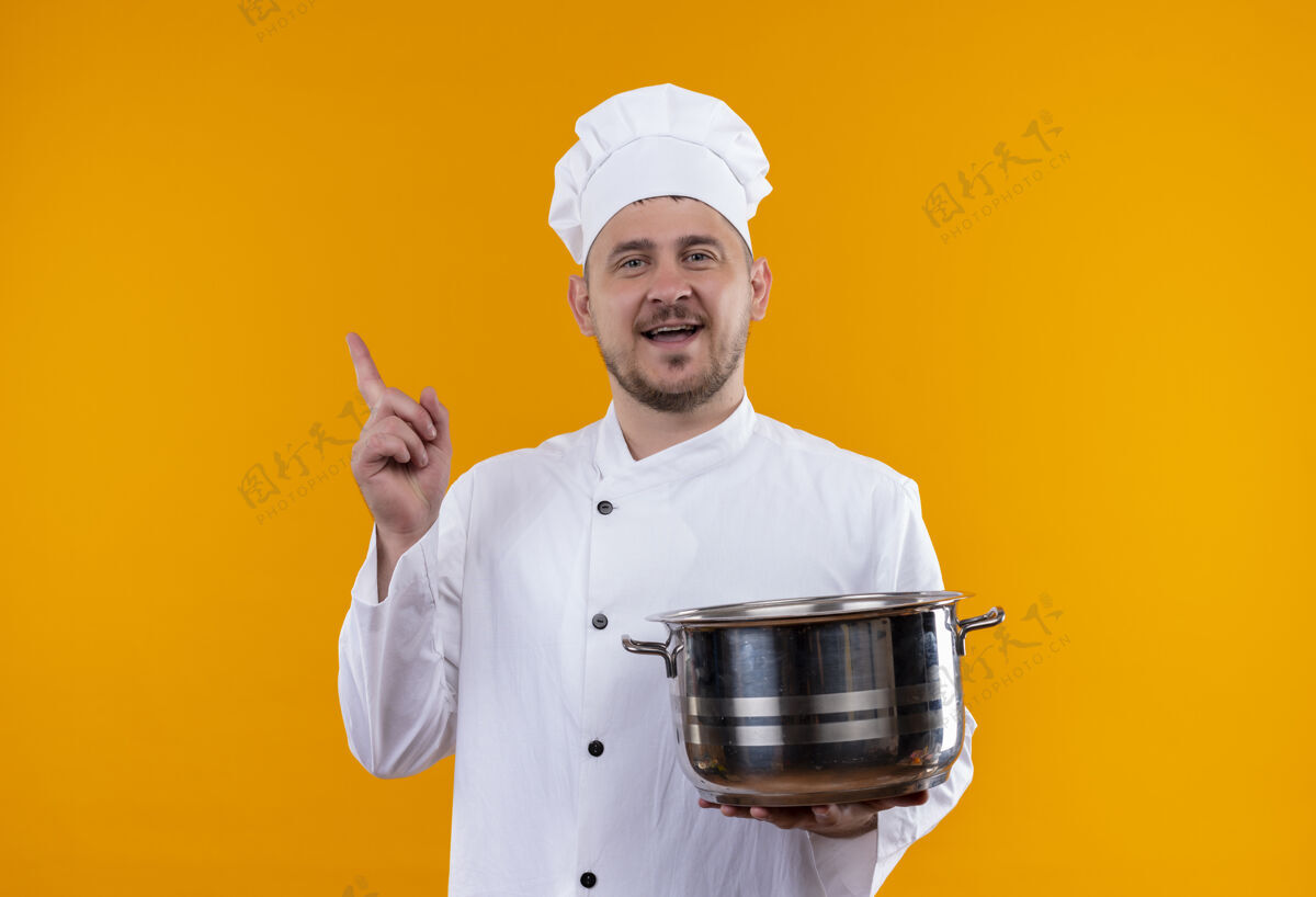 制服快乐的年轻帅哥厨师穿着厨师制服拿着锅 举起手指孤立在橙色的墙上手持快乐厨师