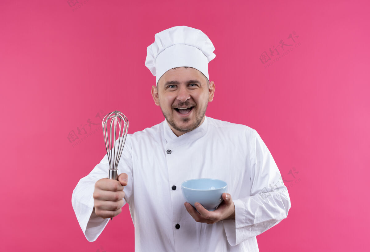 厨师快乐的年轻帅哥厨师穿着厨师制服拿着威士忌和碗隔离在粉红色的墙上碗年轻快乐