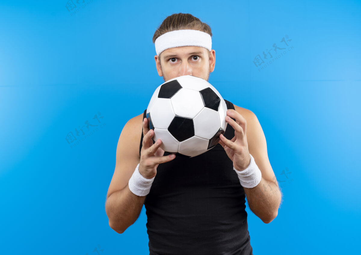 男子令人印象深刻的年轻英俊的运动男子戴着头带和腕带拿着足球 躲在它后面隔离在蓝色的墙上头带年轻持有