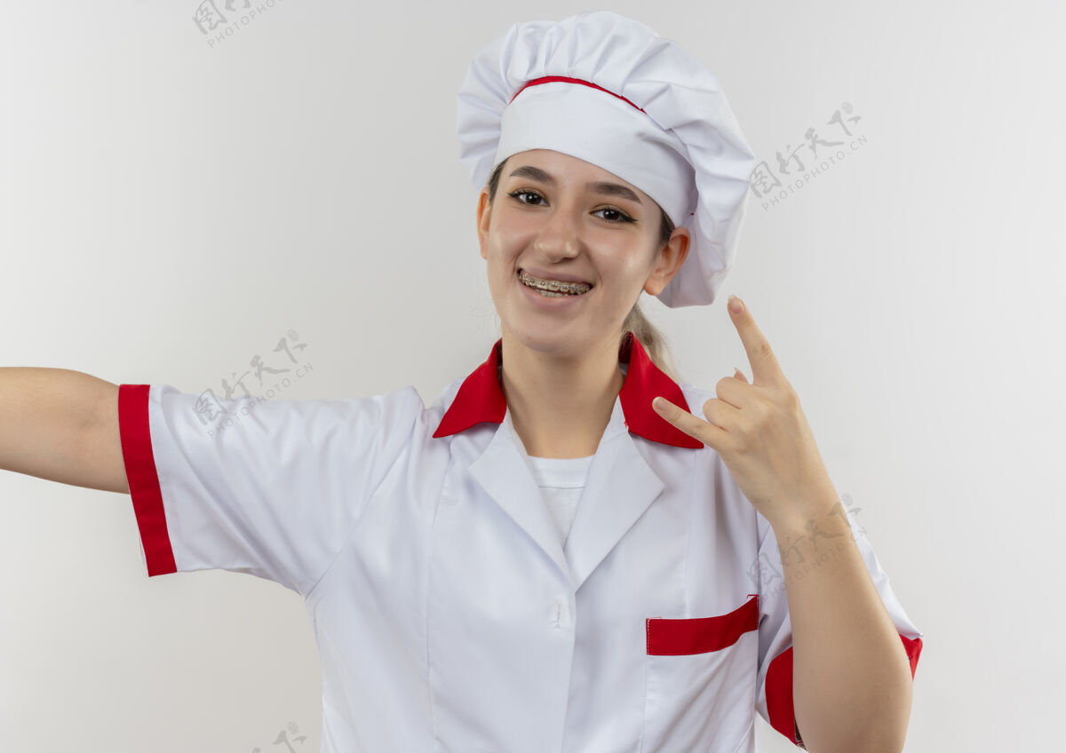 烹饪年轻漂亮的厨师穿着厨师制服 戴着牙套 张开双臂 在孤零零的白墙上做着摇滚的手势牙科年轻手臂