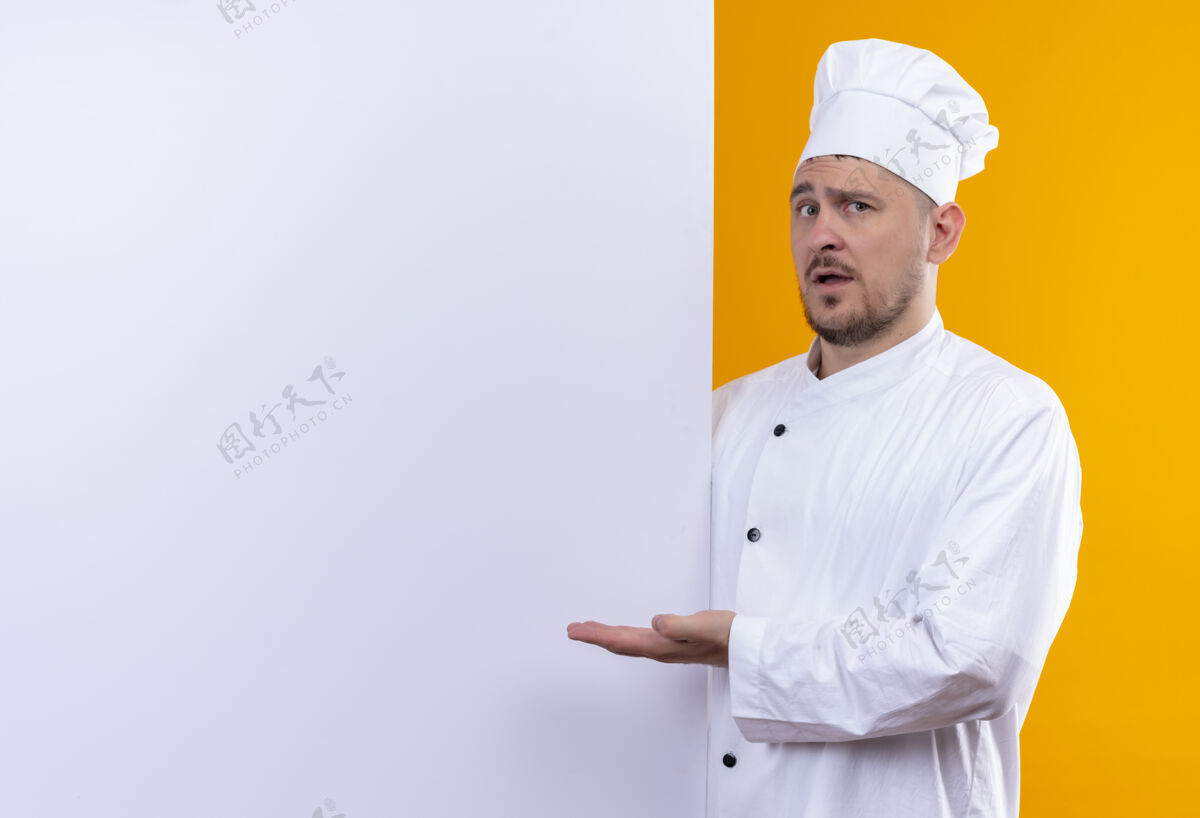 指向穿着厨师制服的年轻帅哥厨师站在白色墙壁后面 用手指着隔离在橙色墙壁上的复制空间烹饪手墙