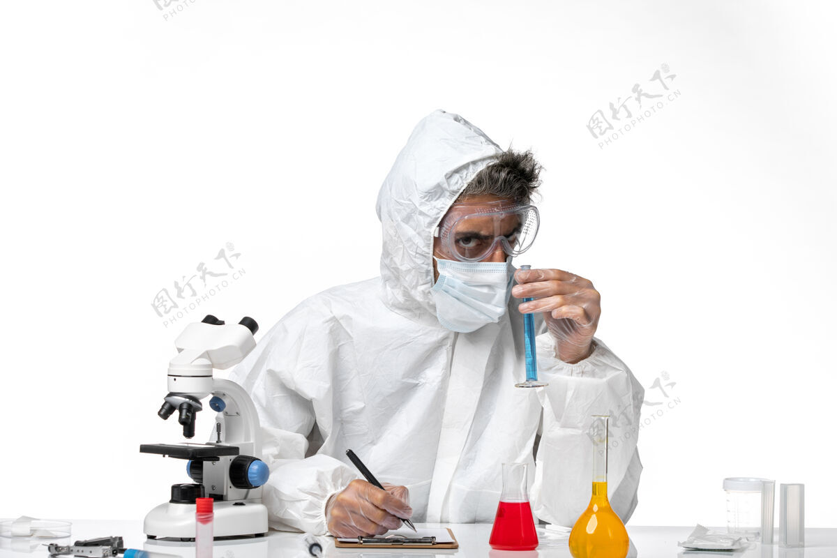 医生穿防护服的男医生写下笔记 拿着蓝色溶液在白色上实验室外套下蓝色