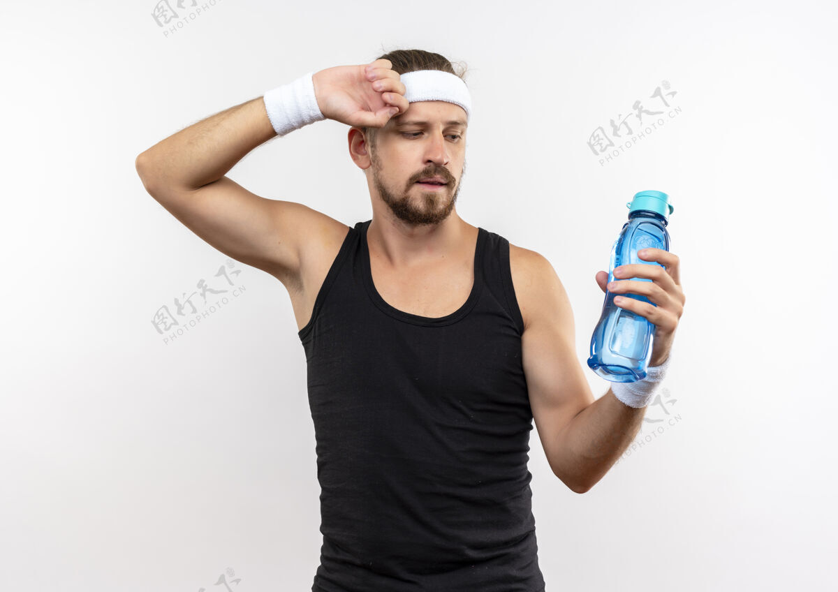 戴自信的年轻帅气的运动型男人戴着头带和腕带 手放在头上看着水瓶 隔离在白色的墙上头带手瓶子