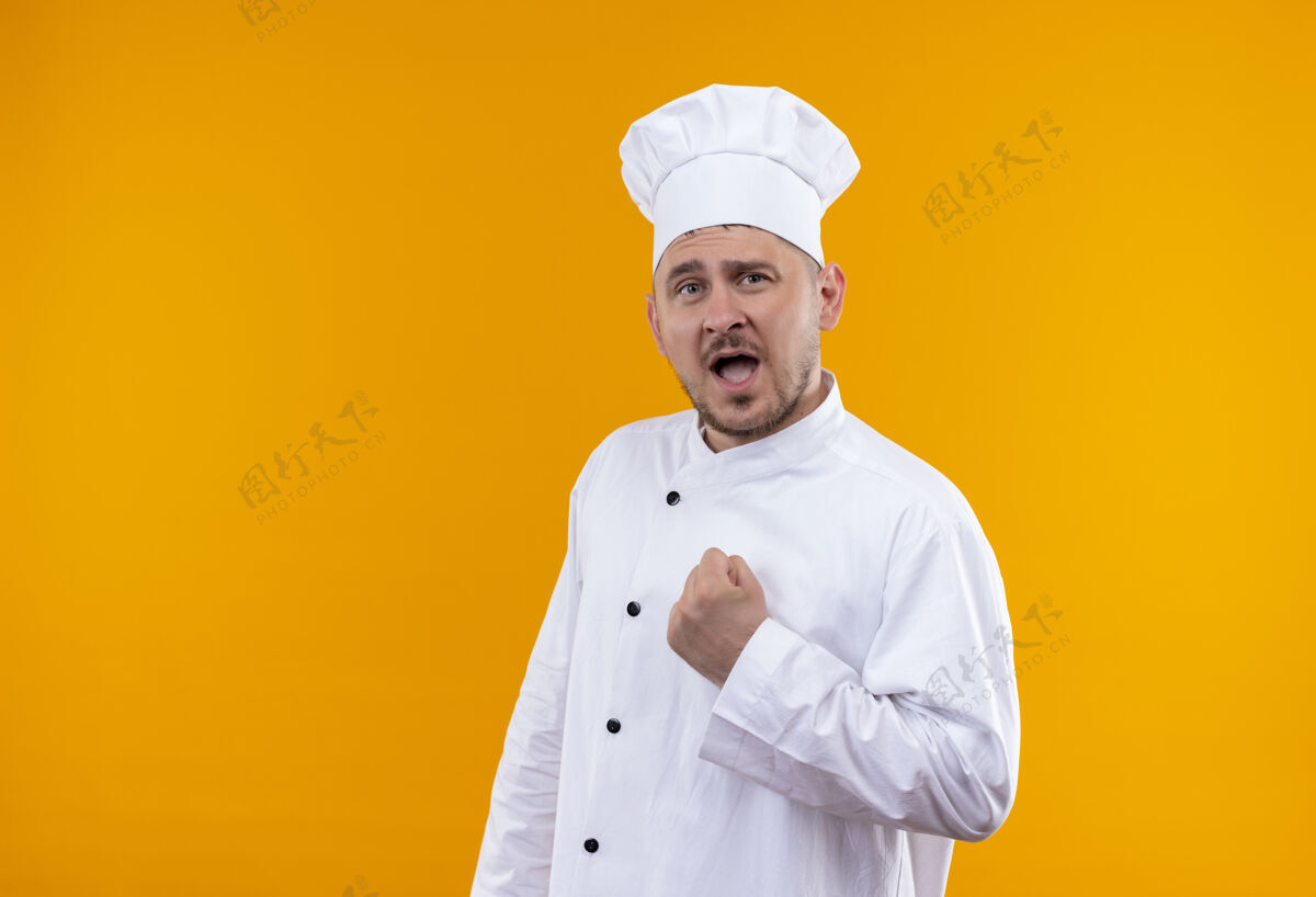 拳头自信的年轻帅哥厨师身着厨师制服 握紧拳头孤立在橙色的墙上握紧厨艺年轻