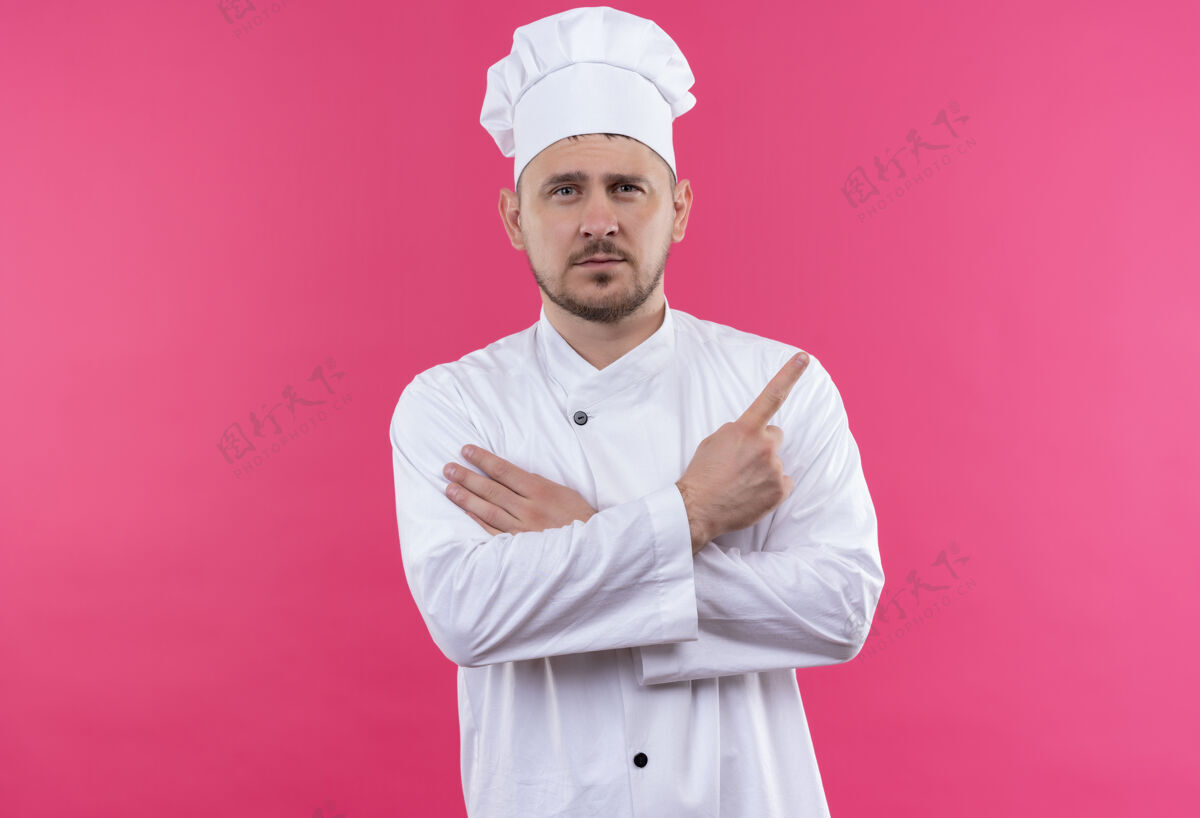 侧身自信的年轻帅哥厨师身着厨师制服 手放在胳膊上 指着粉色墙上孤立的一面手臂自信厨师