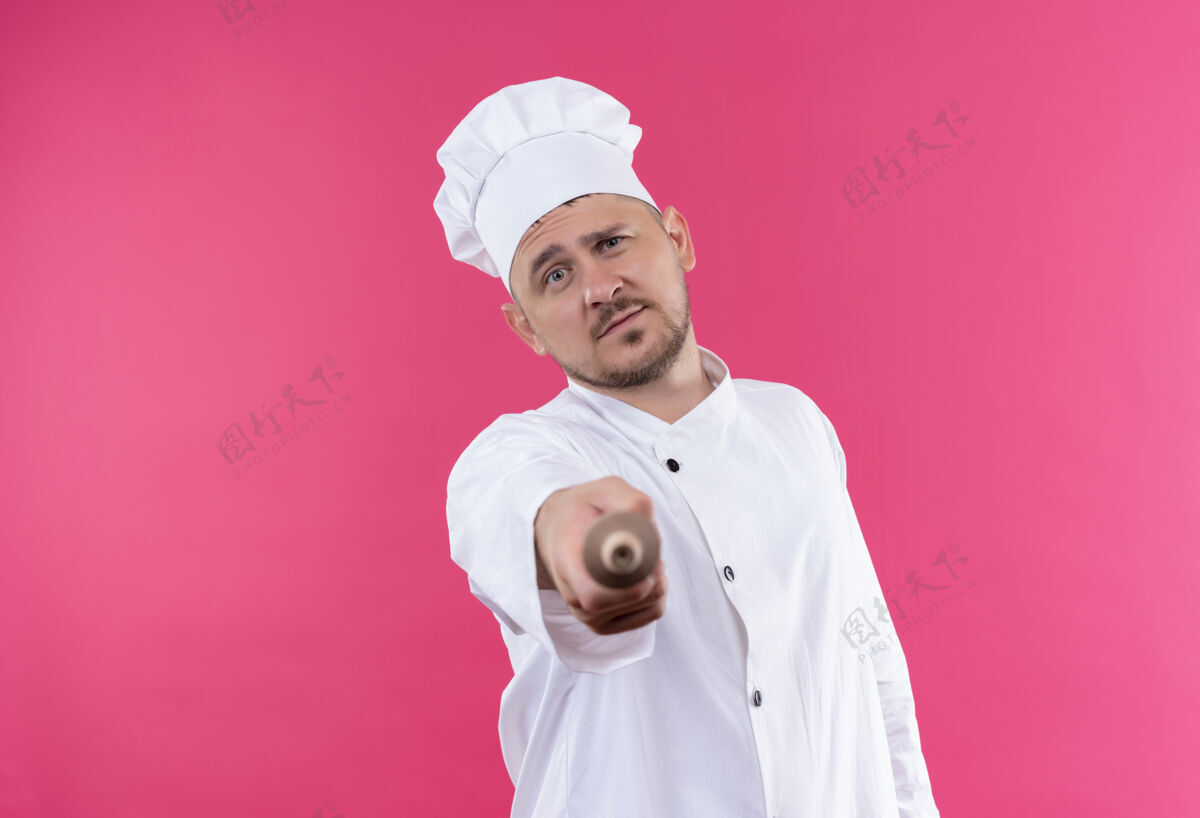 自信自信的年轻帅哥厨师身着厨师制服指着粉色墙上孤立的擀面杖滚制服厨师