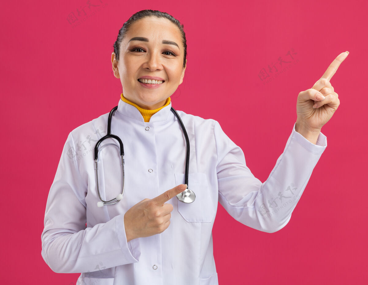 微笑年轻的女医生 身穿白色药衣 脖子上戴着听诊器 脸上带着微笑 食指指向粉色墙壁上的一边手指女人年轻人