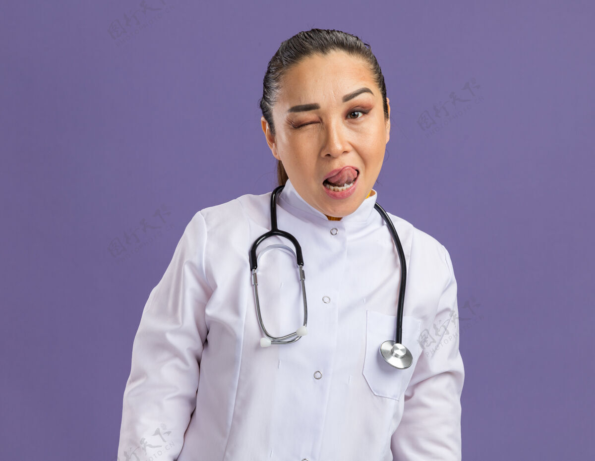 棒快乐的年轻女医生 穿着白药衣 脖子上戴着听诊器 站在紫色的墙上眨眼 伸出舌头女人眨眼外套
