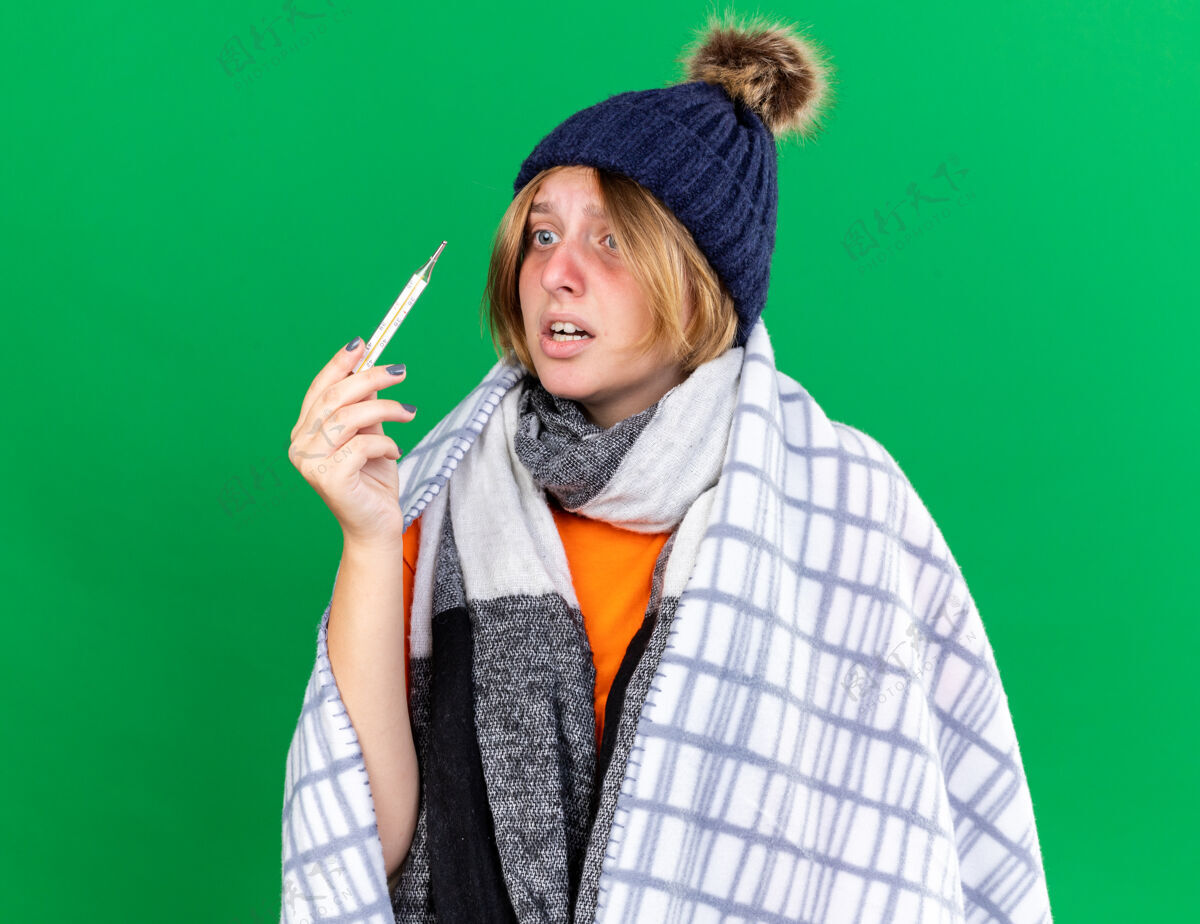 包裹不健康的年轻女子裹着毯子 戴着帽子 用体温计测量体温 患流感 发烧 站在绿色的墙上 看起来很担心不健康痛苦发烧