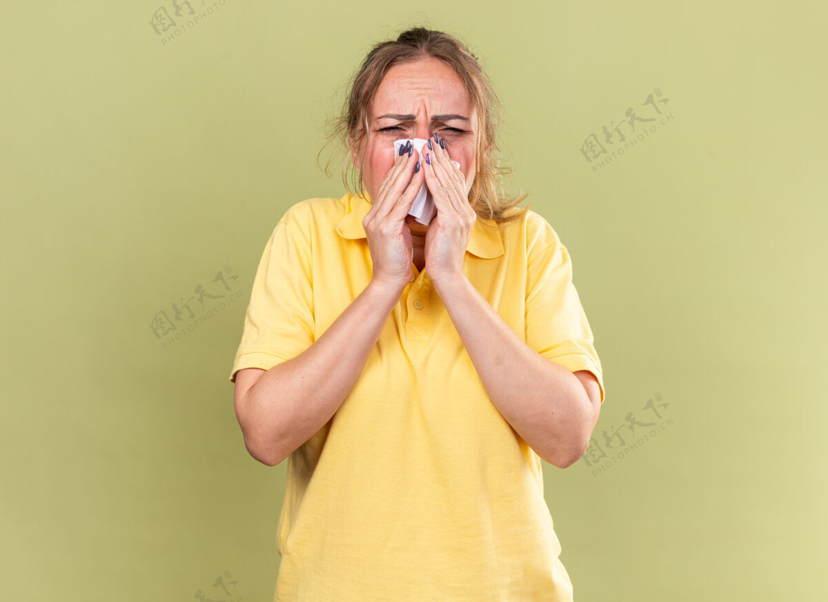 打喷嚏穿着黄色衬衫的不健康的女人感觉很可怕 患流感和感冒 流鼻涕 打喷嚏 用纸巾站在绿色的墙上跑步女人不健康