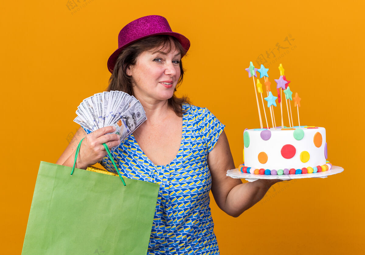 中间快乐的中年妇女戴着派对帽 手里拿着带礼物的纸袋 手里拿着生日蛋糕和现金 微笑着站在橙色的墙上庆祝生日派对女人庆祝现金