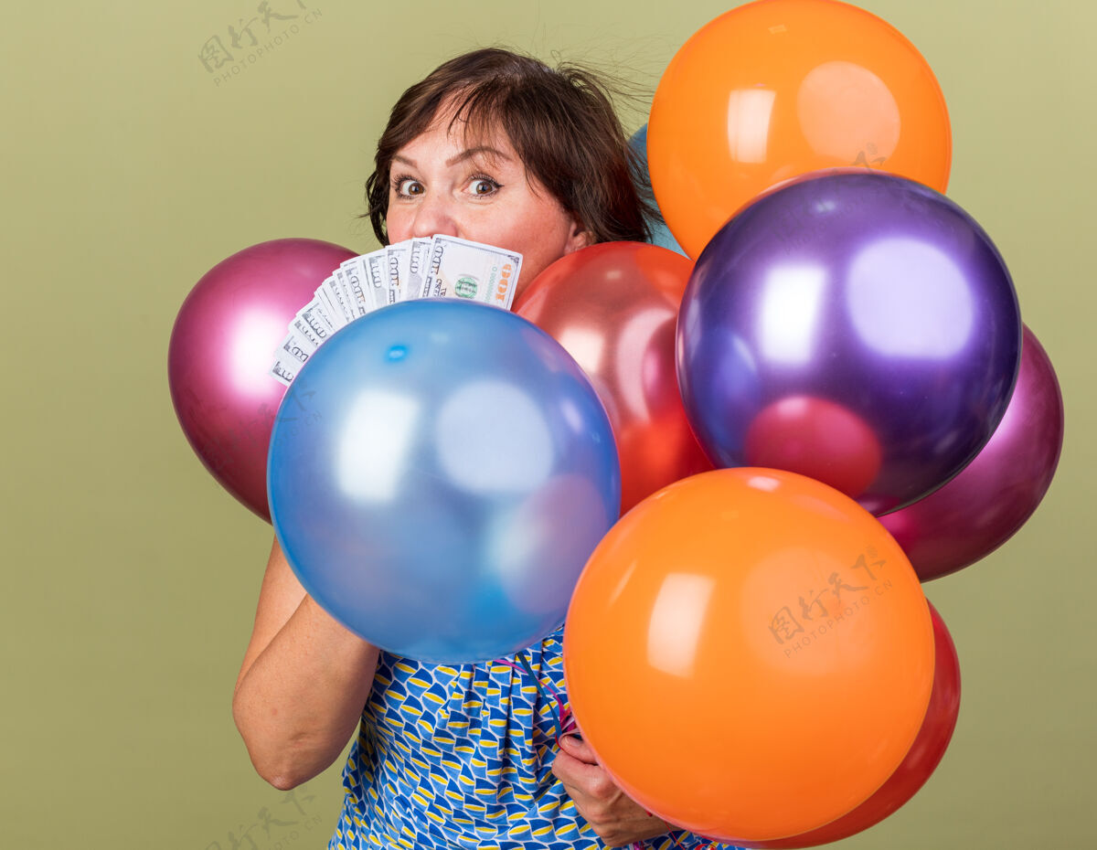 束中年妇女拿着一堆五颜六色的气球 手里拿着现金 站在绿色的墙上惊喜地庆祝生日派对年龄立场女人