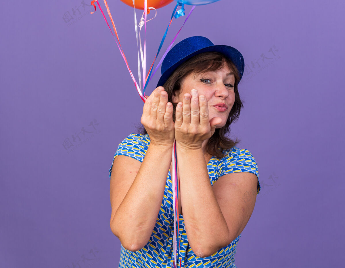吻快乐开朗的中年妇女戴着派对帽 手持一束五颜六色的气球 站在紫色的墙上亲吻庆祝生日派对女人吹束