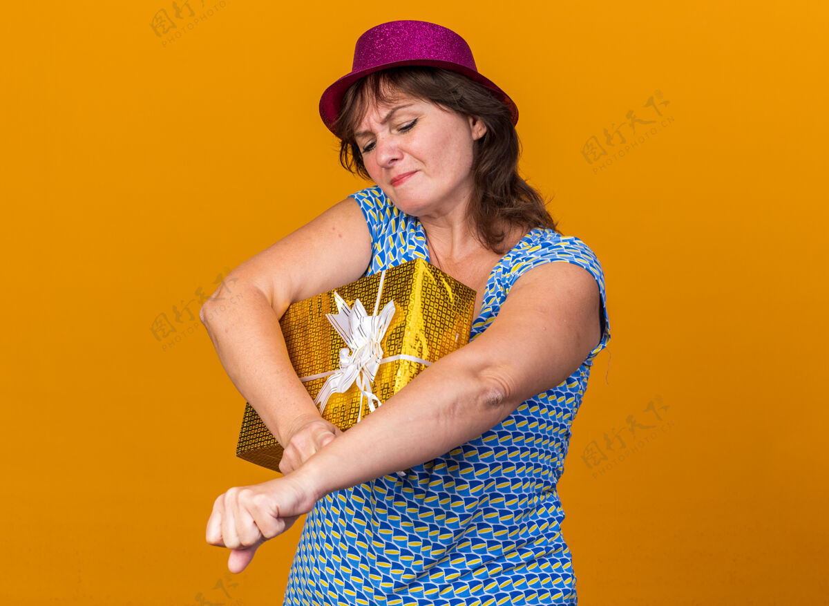 女人中年妇女戴着派对帽 手里拿着礼物 神情困惑 不高兴地站在橘色的墙上庆祝生日派对庆祝年龄立场