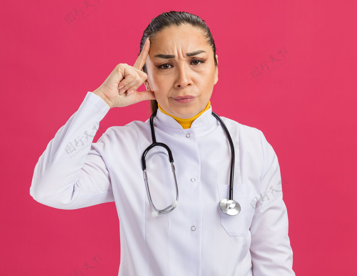 表情年轻的女医生 穿着白药衣 脖子上戴着听诊器 用食指指着太阳穴 脸上带着怀疑的表情站在粉红色的墙上女人围着怀疑