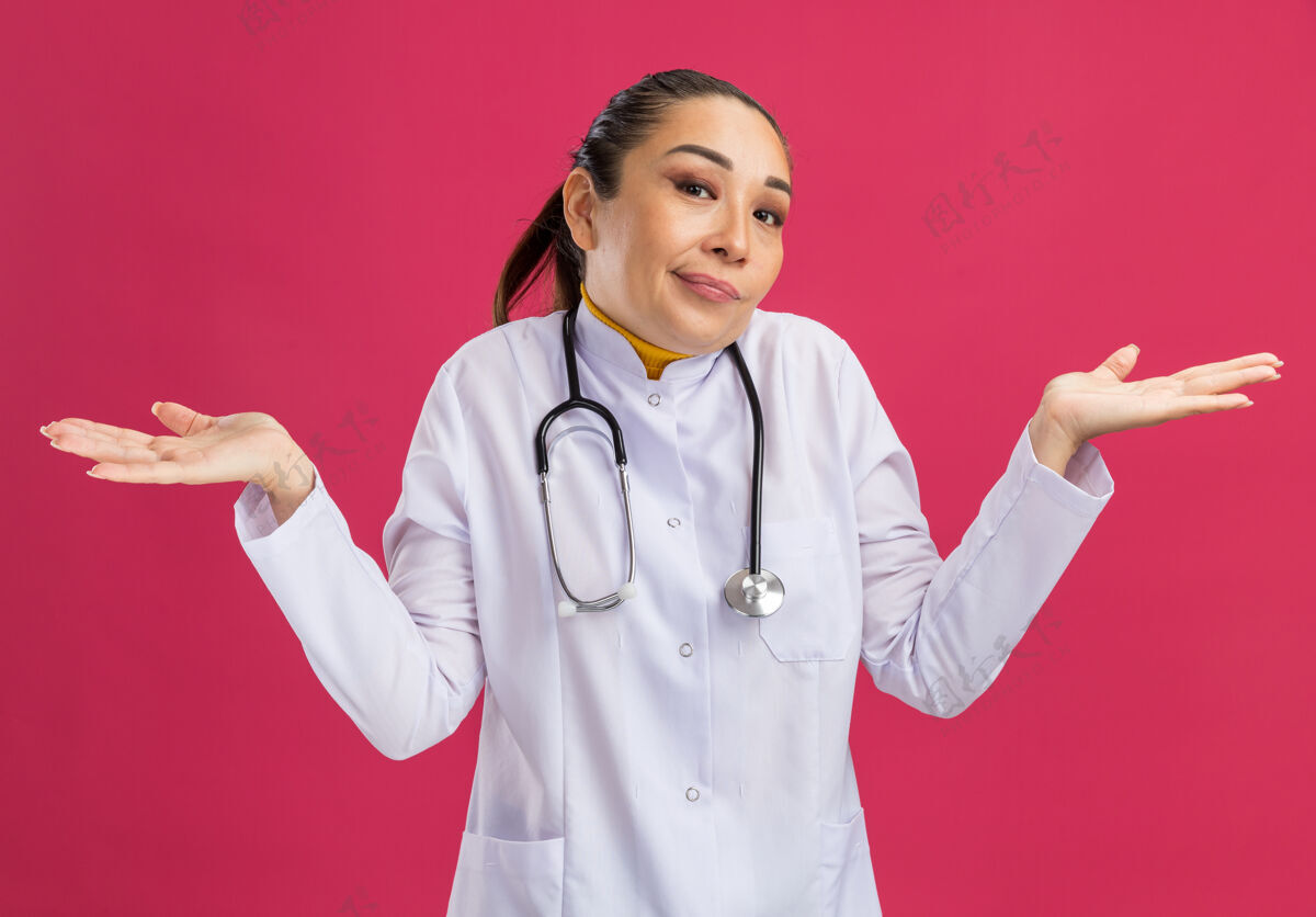 外套年轻的女医生穿着白药衣 脖子上戴着听诊器 站在粉红色的墙上 困惑地耸着肩膀 没有回答年轻药物站