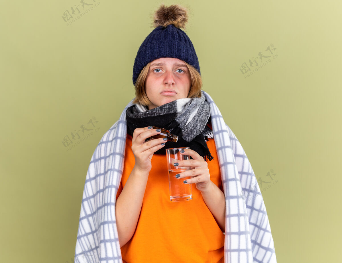 感觉不健康的年轻女子 戴着暖和的帽子 脖子上围着围巾 裹着毯子 感觉不舒服 滴在一杯患流感的水里 站在绿色的墙上滴帽子不健康