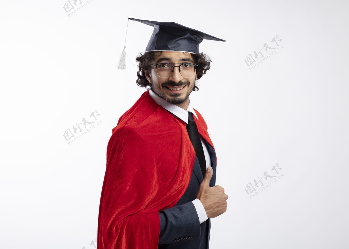 侧年轻的高加索超级英雄面带微笑 戴着眼镜 身穿西装 披着红色斗篷 头戴毕业帽 站在一旁竖起大拇指超级英雄向上帽子