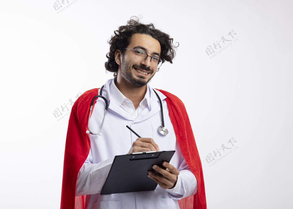 红年轻的高加索超级英雄微笑着 戴着眼镜 身穿医生制服 披着红色斗篷 脖子上戴着听诊器 手里拿着铅笔和剪贴板听诊器光学微笑