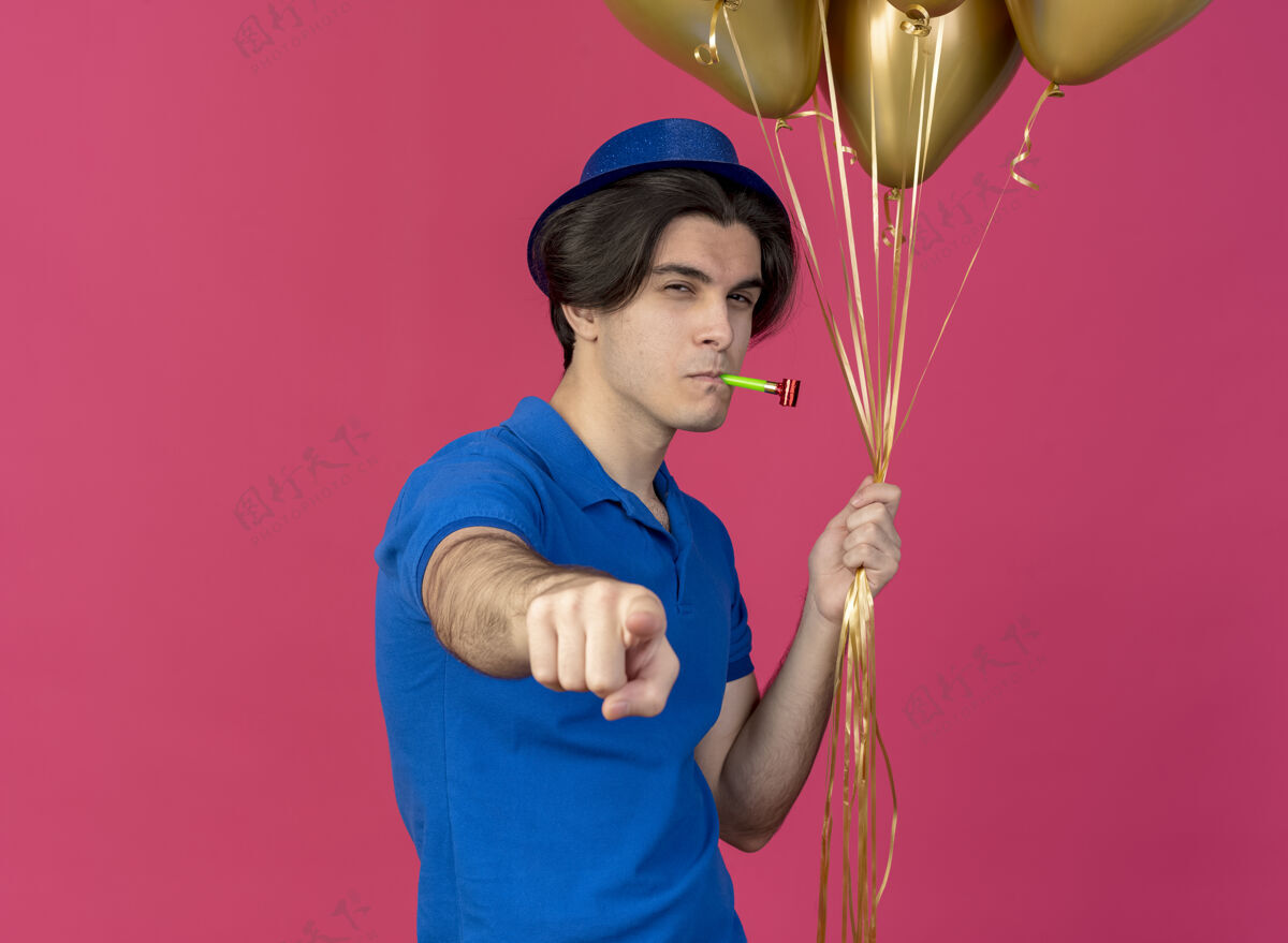 派对自信英俊的白人男子戴着蓝色的派对帽手持氦气球吹着派对哨子指着摄像机指向穿氦
