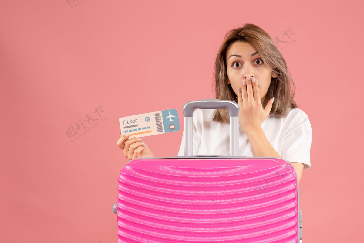人在粉红色手提箱后面拿着票的年轻女子感到惊奇模型手提箱可爱