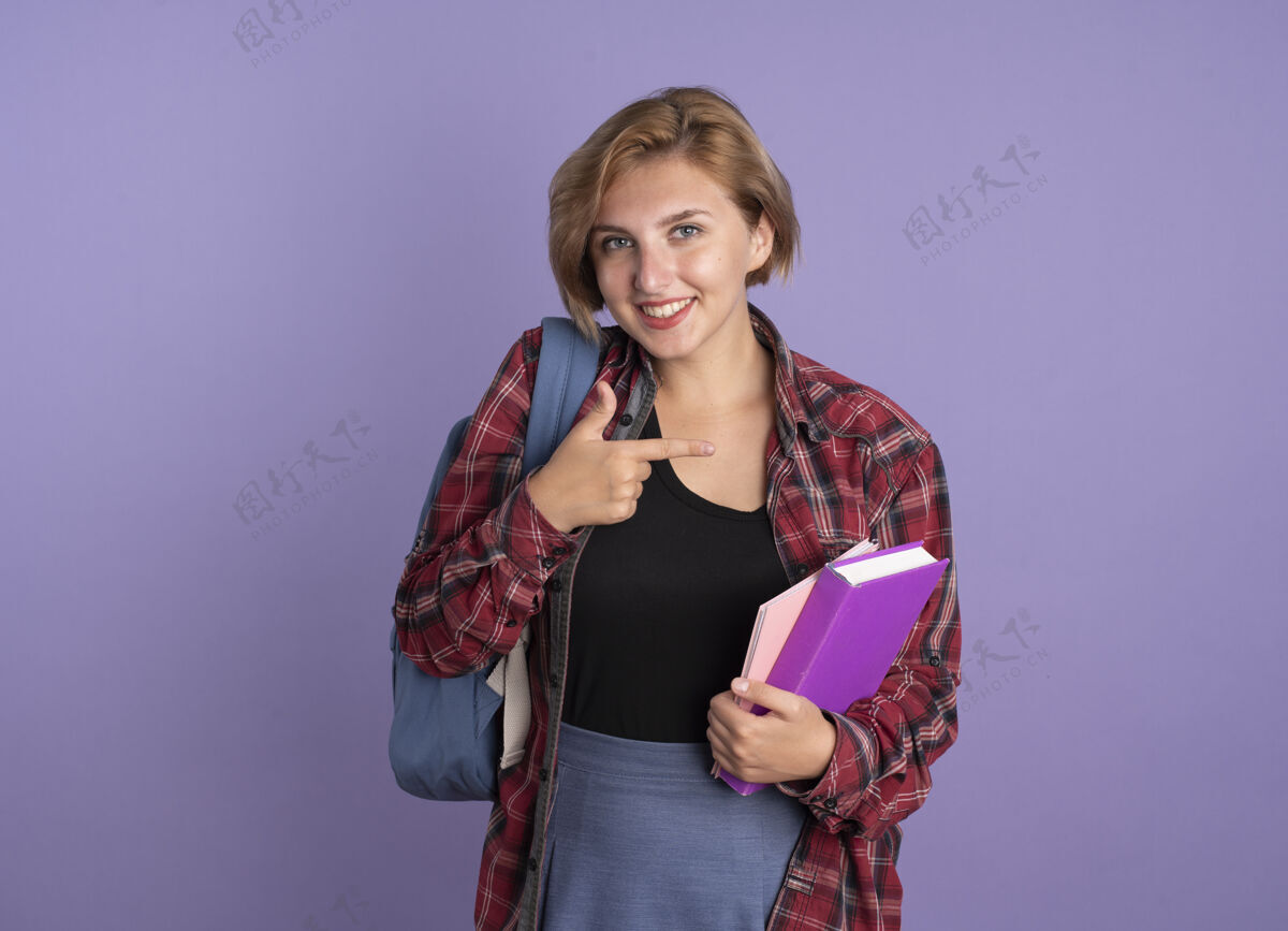 穿面带微笑的年轻斯拉夫女学生背着背包拿着书和笔记本指着身边年轻指向紫色